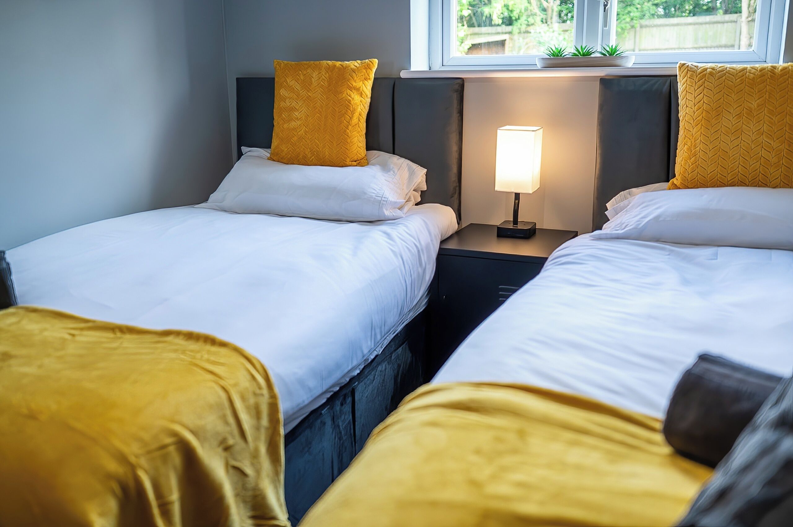 A hotel room with two single beds, each with a headboard, a bedside table with a lamp, and a window with a view of greenery outside.
