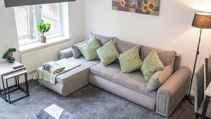 Een woonkamer met een grijze bank, groene kussens, een bijzettafeltje met een plant, een radiator en een schilderij van paarden.