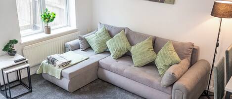A living room with a grey sofa, green cushions, a small table with a plant, a radiator, and a painting of horses.