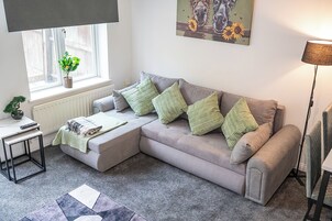 A living room with a grey sofa, green cushions, a small table with a plant, a radiator, and a painting of horses.