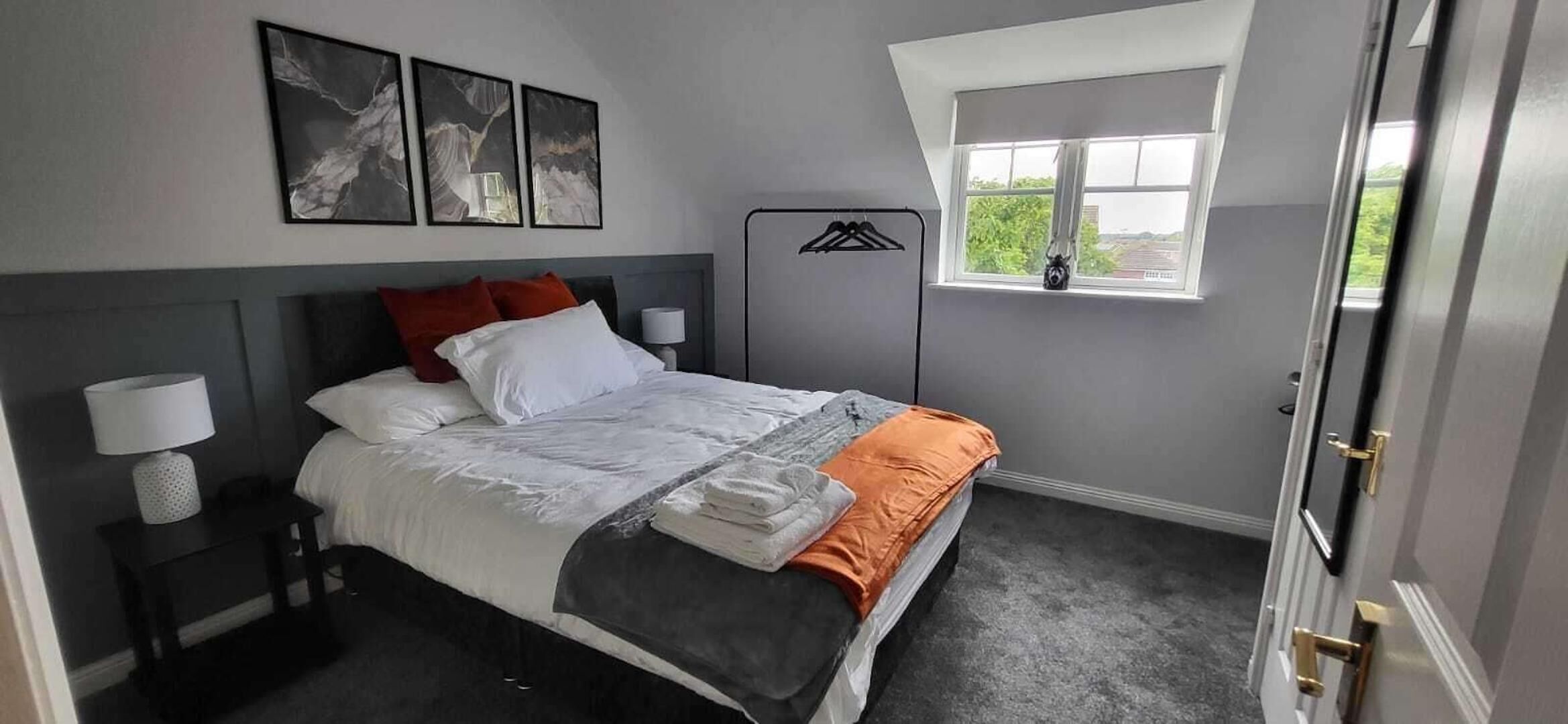 Un dormitorio con cama, mesitas de noche, ventana y cuadros en la pared.