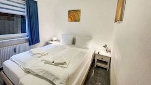 A hotel room with a double bed, two bedside tables, a nightstand, a window with curtains, a radiator, and a painting on the wall. - Lovely apartment for 2 people with WIFI, TV, patio and pets allowed (Westerland)