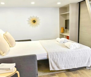 Quarto de hotel com uma cama, uma mesa de cabeceira com um livro, uma luminária em forma de sol na parede e um guarda-roupa.