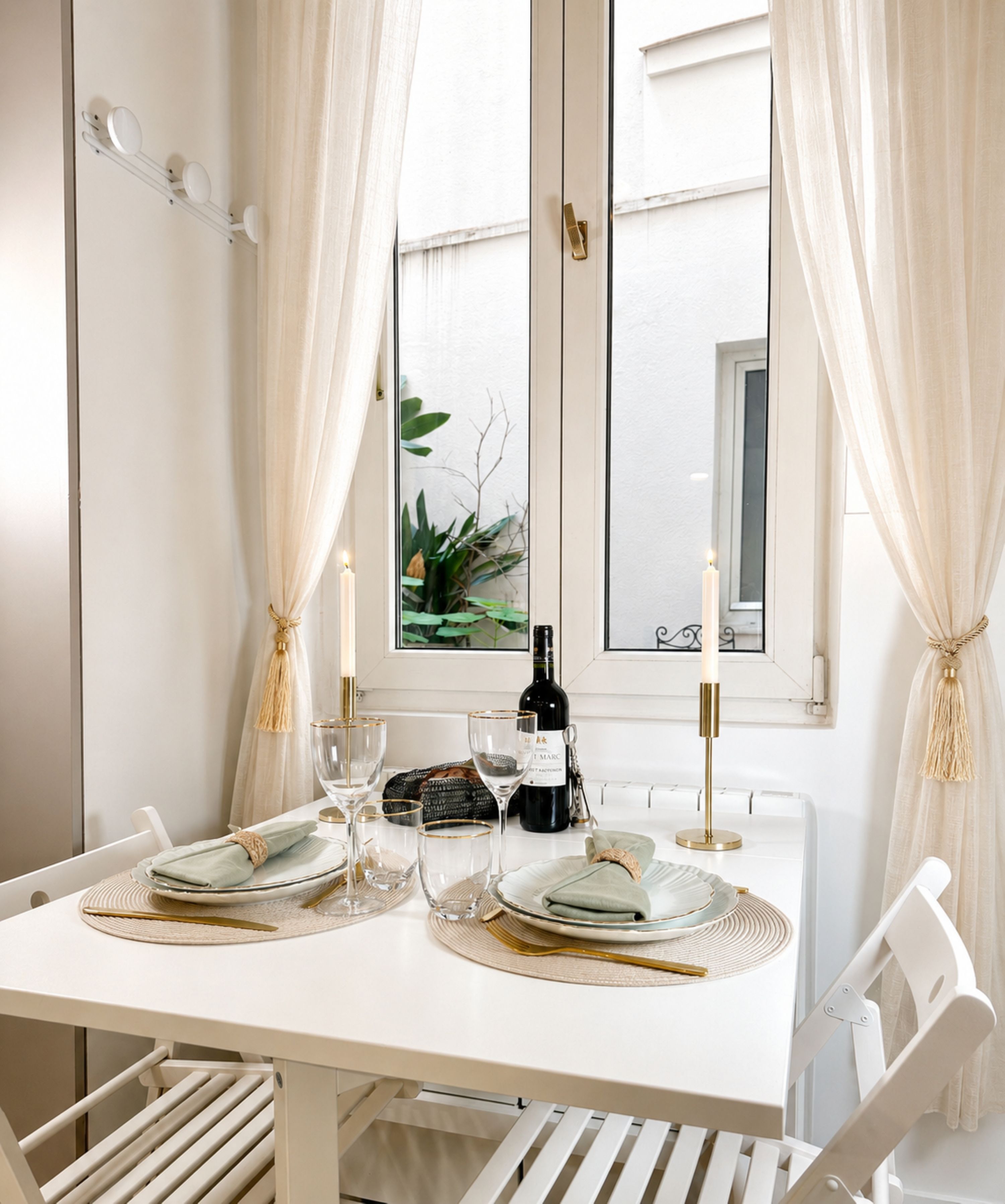A dining table set with plates, glasses, and a bottle, with white curtains and a window view.