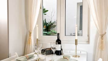 A dining table set with plates, glasses, and a bottle, with white curtains and a window view.