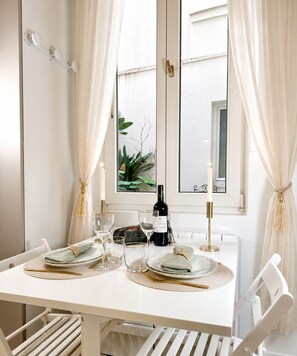 A dining table set with plates, glasses, and a bottle, with white curtains and a window view. - 1571 Lombards (Paris)