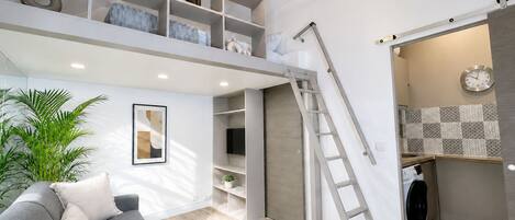 A modern living space with a grey sofa, a coffee table, a ladder, and a built-in washer and dryer.