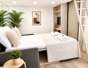 Een moderne hotelkamer met een bed, een bureau, een stoel, een potplant, een ingelijst kunstwerk en een televisie op een plank.