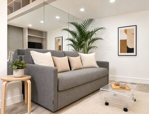 A modern living room with a grey sofa, a small side table with a plant, a glass coffee table with a tray, and a large potted plant. - 1571 Lombards (Paris)