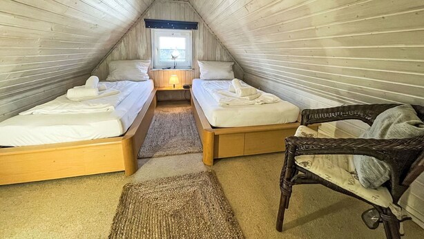 A cozy attic bedroom with two beds, a window, a lamp, and a wicker chair.