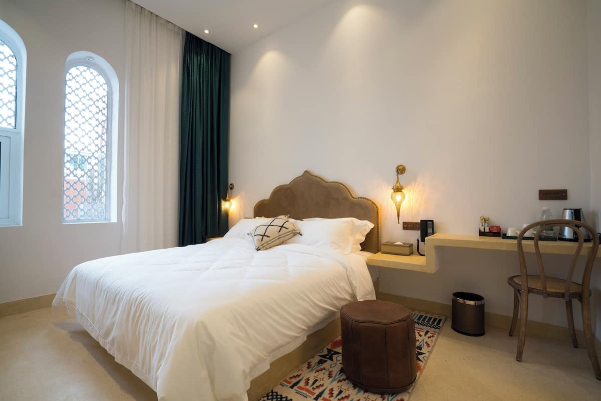 Una cama bien hecha con cabecera, lámparas de noche, una mesita, una silla y una ventana con cortinas.