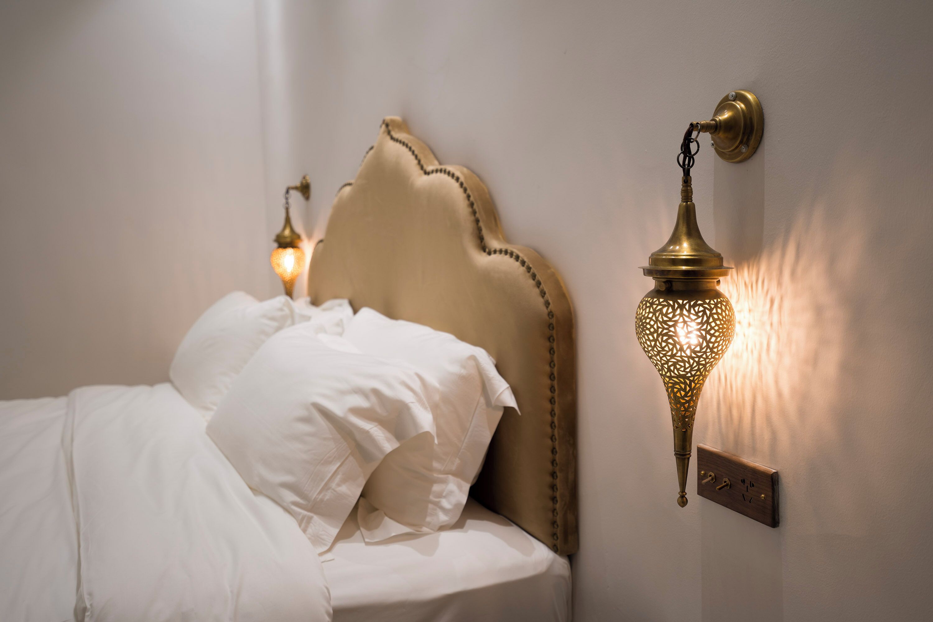 Una cama con sábanas blancas y un cabecero decorativo, una lámpara de pared y un panel de control.