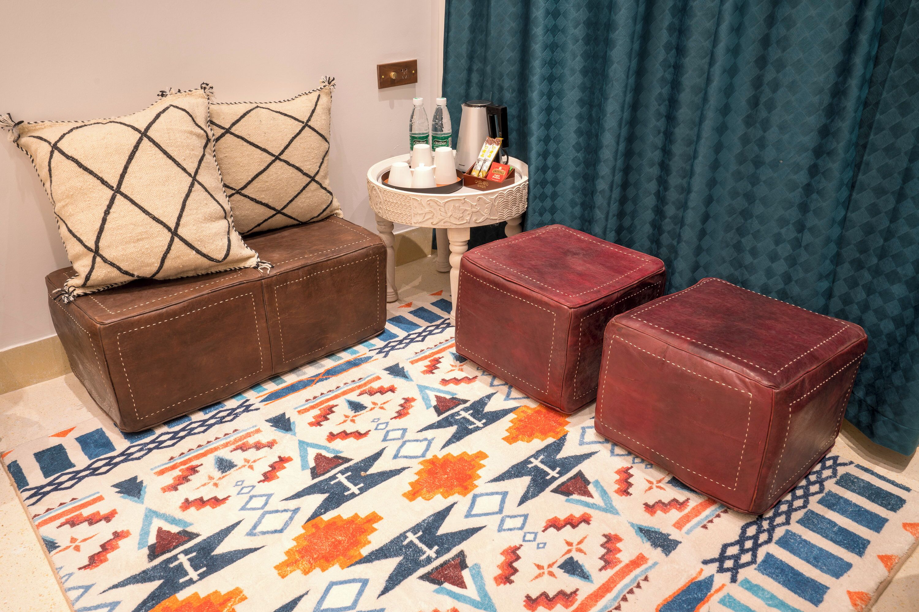 A room with a brown leather sofa, two red leather ottomans, a patterned rug, a small round table with a coffee maker and bottles, and a blue curtain.