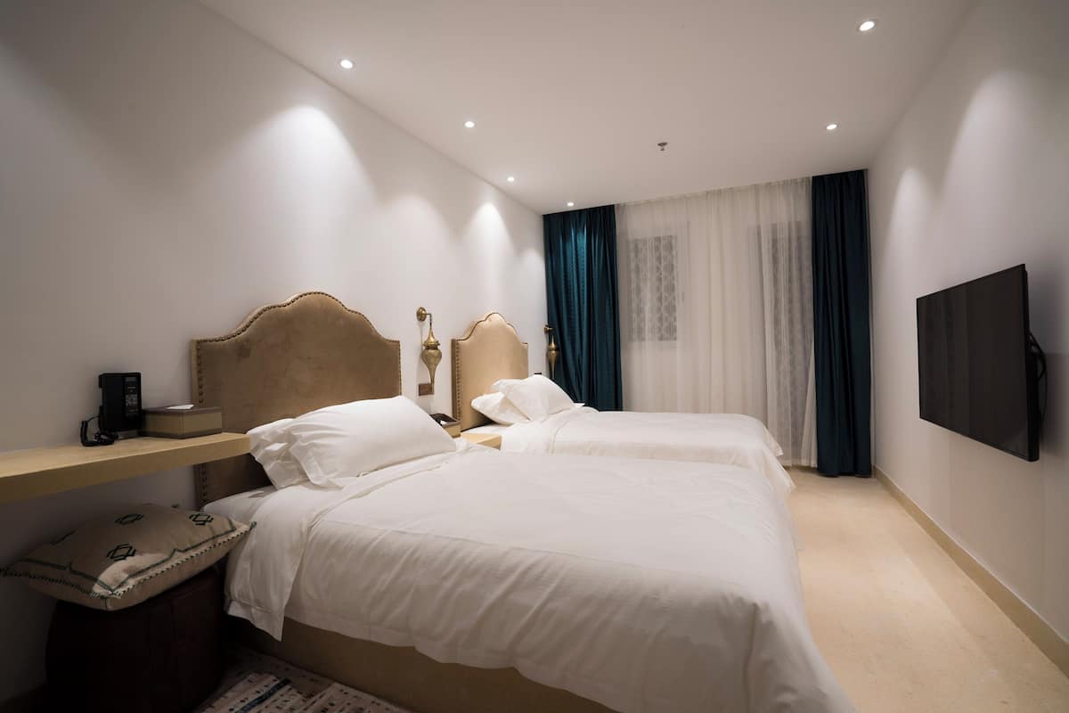 Habitación de hotel con dos camas, una mesita de noche con lámpara, un televisor de pantalla plana montado en la pared y una ventana con cortinas traslúcidas.