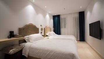 Quarto de hotel com duas camas, uma mesa de cabeceira com abajur, televisão de tela plana na parede e janela com cortinas finas.