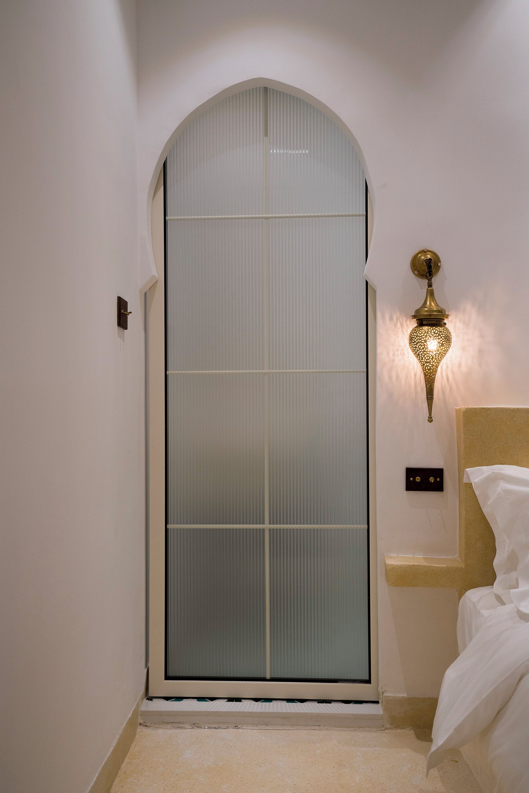 Una habitación con puerta corredera, cama con ropa blanca, lámpara de pared y un estante pequeño.