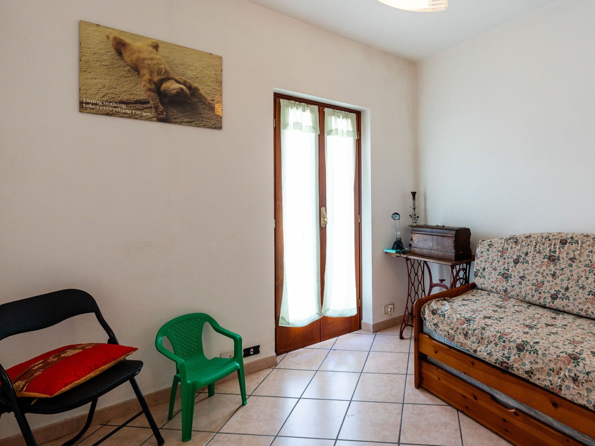 A room with a wooden bed, a green chair, a black chair, a small table with a radio, and a painting of a bear on the wall.
