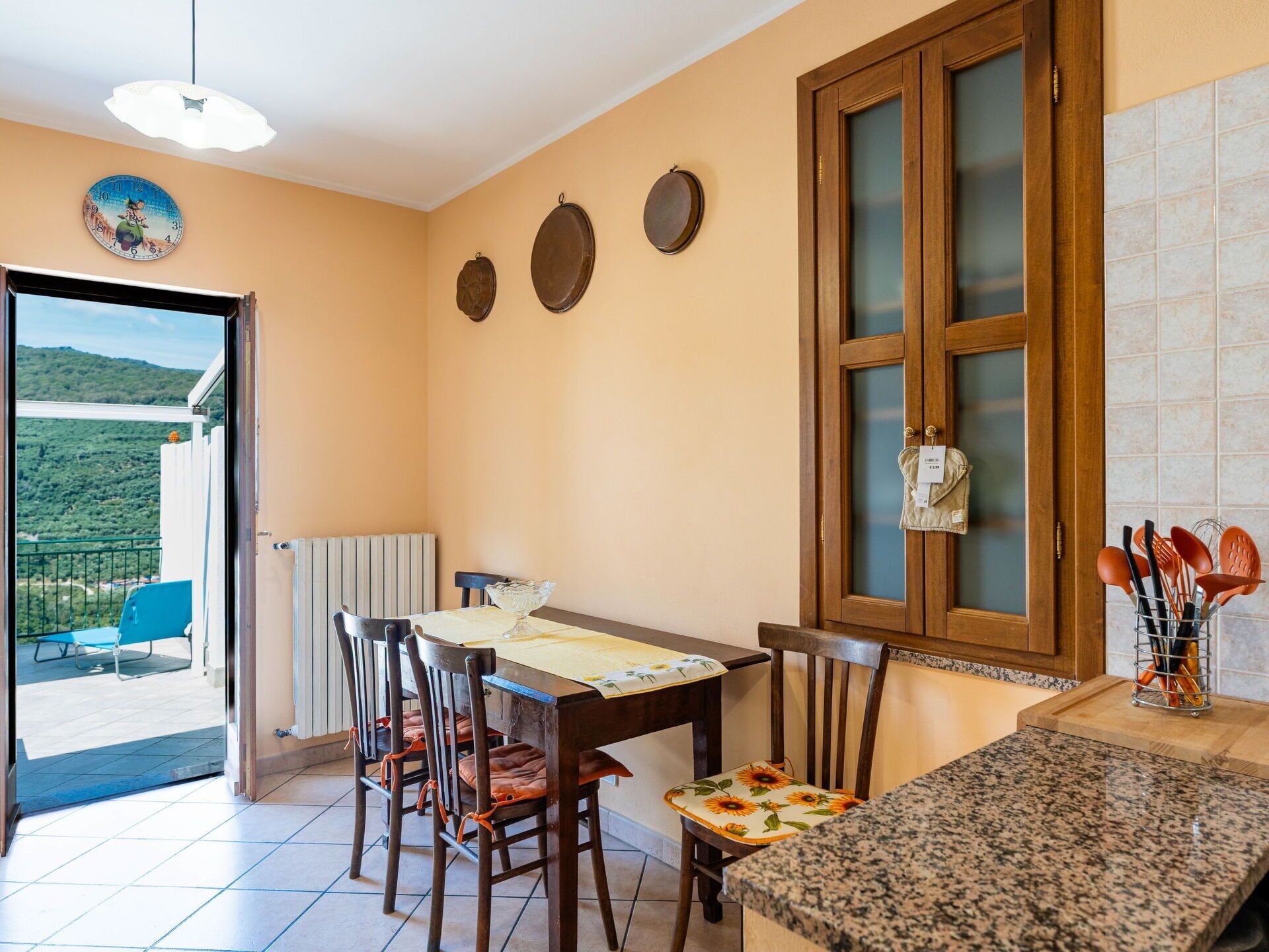 A dining area with a table and chairs, a kitchen counter, and a view of a balcony with a chair.