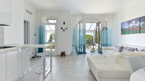 A modern, well-lit living room with a white sofa, a dining table, and a kitchen area with white cabinets. - Beautiful  villa for 6 people with A/C, WIFI, terrace and pets allowed (Castiglione Della Pescaia)
