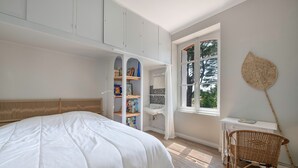 A bedroom with a large bed, a white desk, a wicker chair, a bookshelf, and a window with a view of trees. - Charming villa 250m from beach – sleeps 14 (Saint-Michel-Chef-Chef)