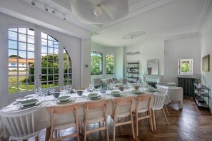 Un coin repas avec une longue table dressée pour le repas, de grandes fenêtres offrant une vue sur l’extérieur et une cheminée en arrière-plan.