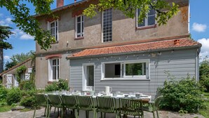 A dining area with a table set for four, surrounded by green chairs, in front of a grey building with a tiled roof. - Charming villa 250m from beach – sleeps 14 (Saint-Michel-Chef-Chef)
