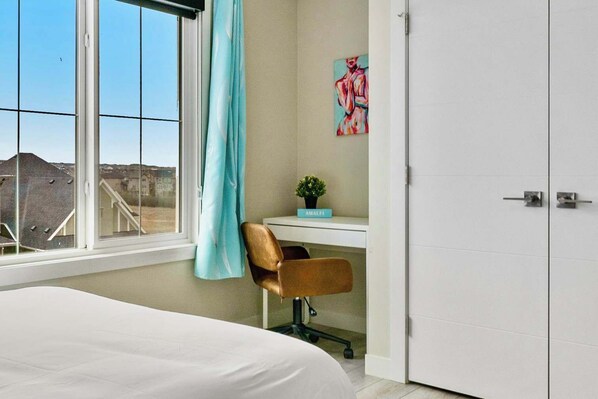 A bedroom with a bed, a desk, and a chair. There is a window with a view of buildings outside.