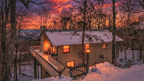 Un chalet confortable avec une terrasse en bois, entouré d’un paysage enneigé et d’arbres, sous un ciel flamboyant au coucher du soleil.