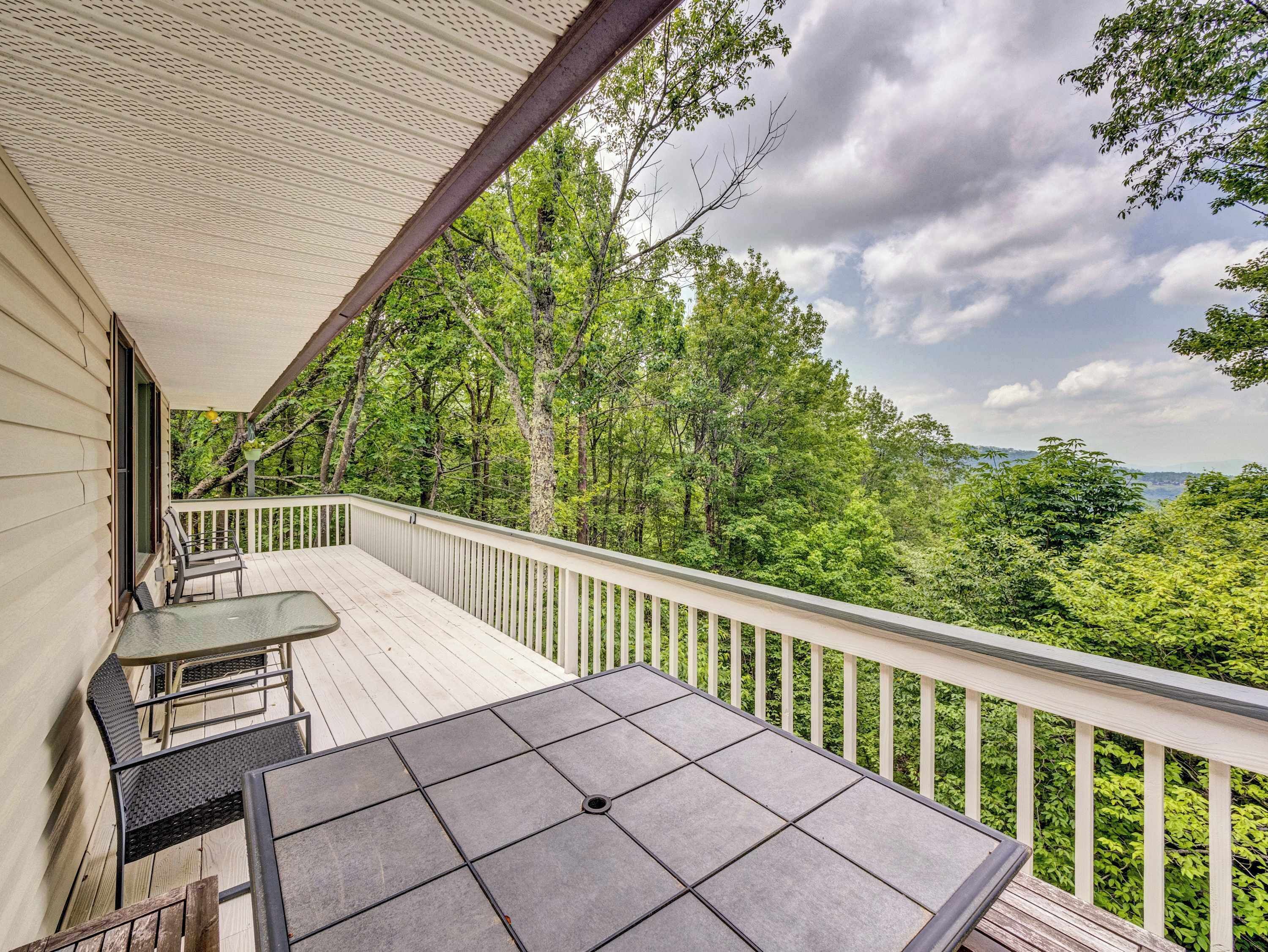 A covered outdoor deck with a table and chairs, surrounded by a white railing and lush greenery.