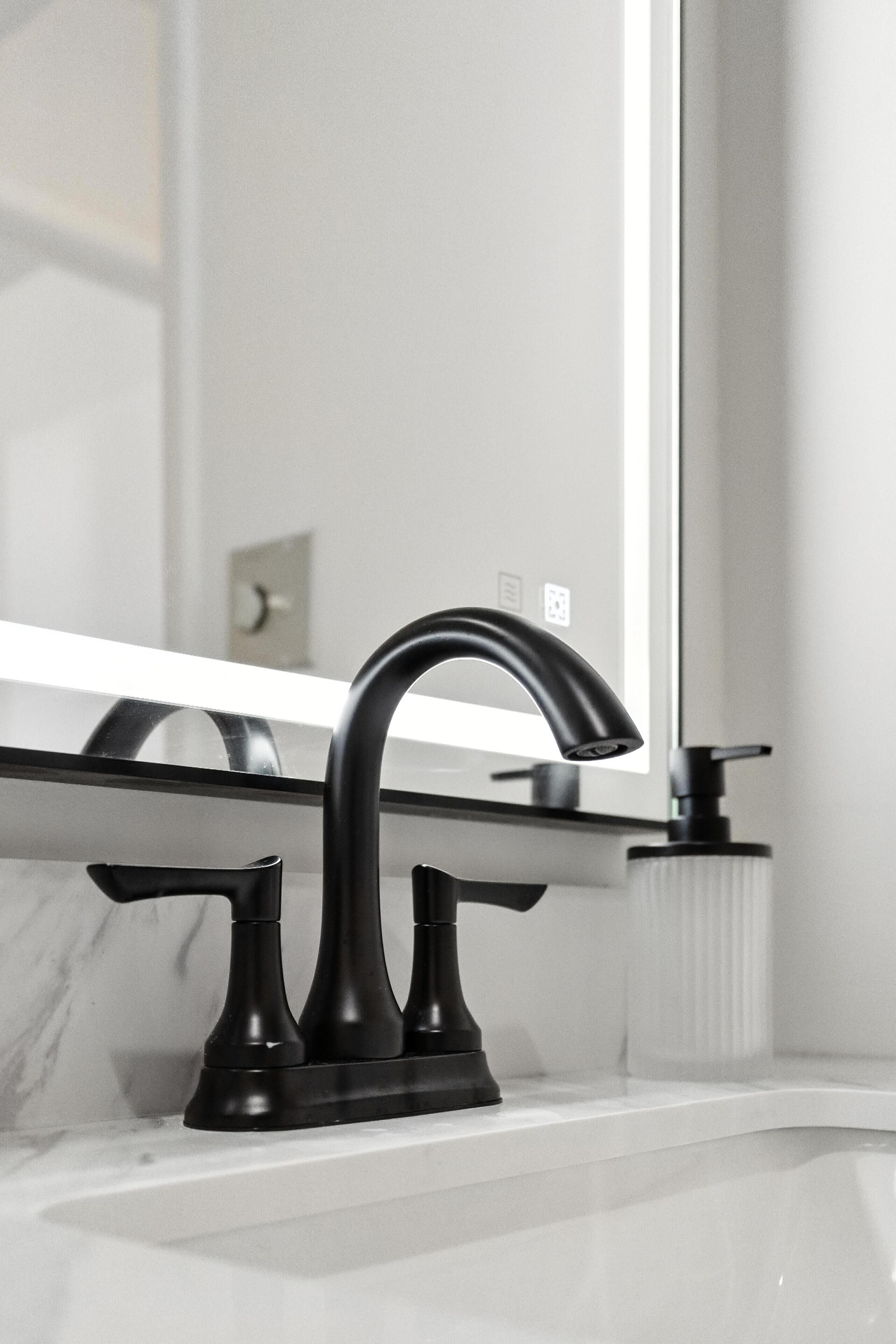 A modern bathroom with a black faucet, a soap dispenser, and a mirror reflecting a light switch and outlet.