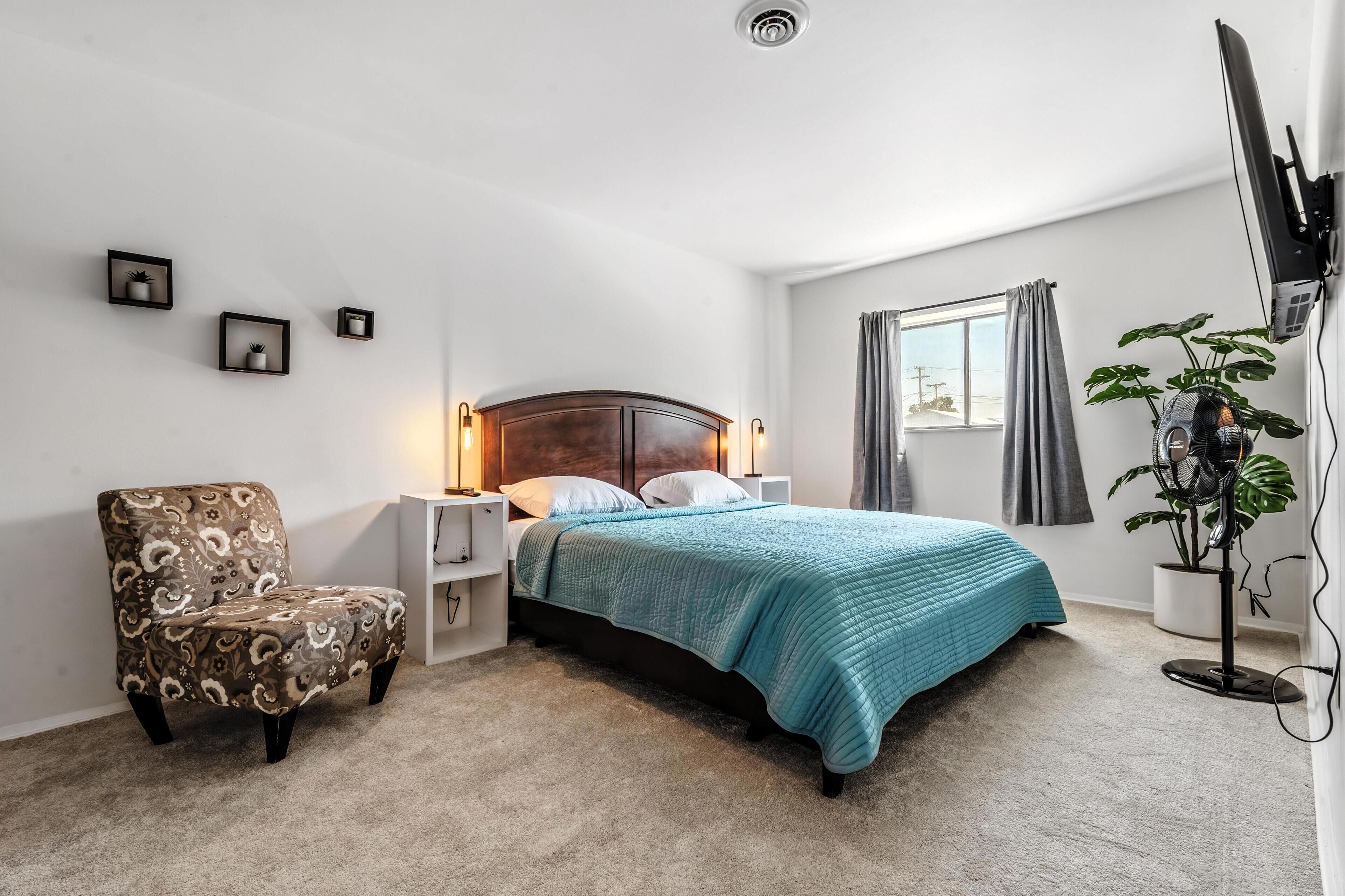 A bedroom with a bed, a chair, bedside tables, wall-mounted lights, a window with curtains, and a potted plant.