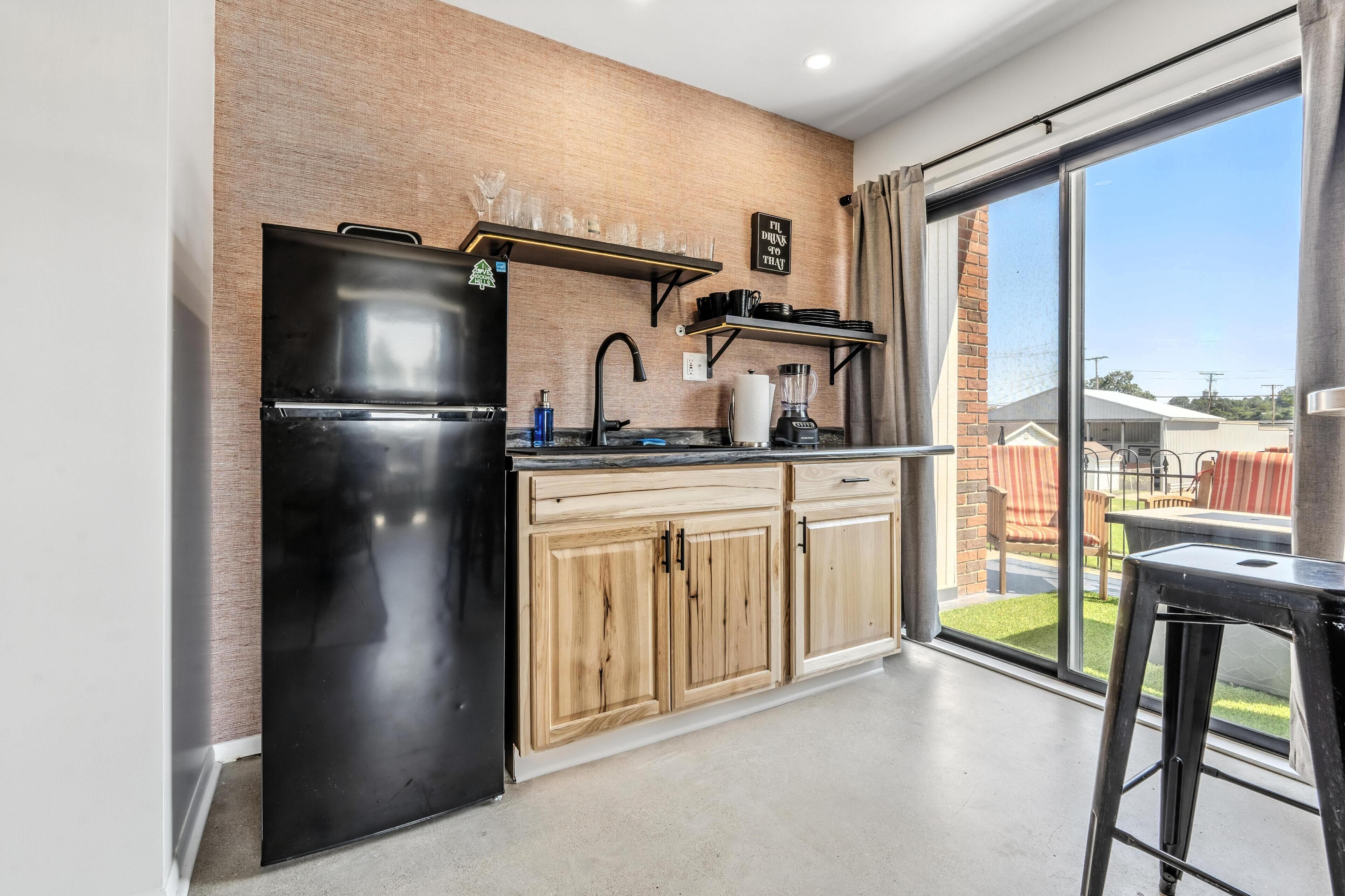 A modern kitchen with a black refrigerator, wooden cabinets, and a window overlooking a backyard.