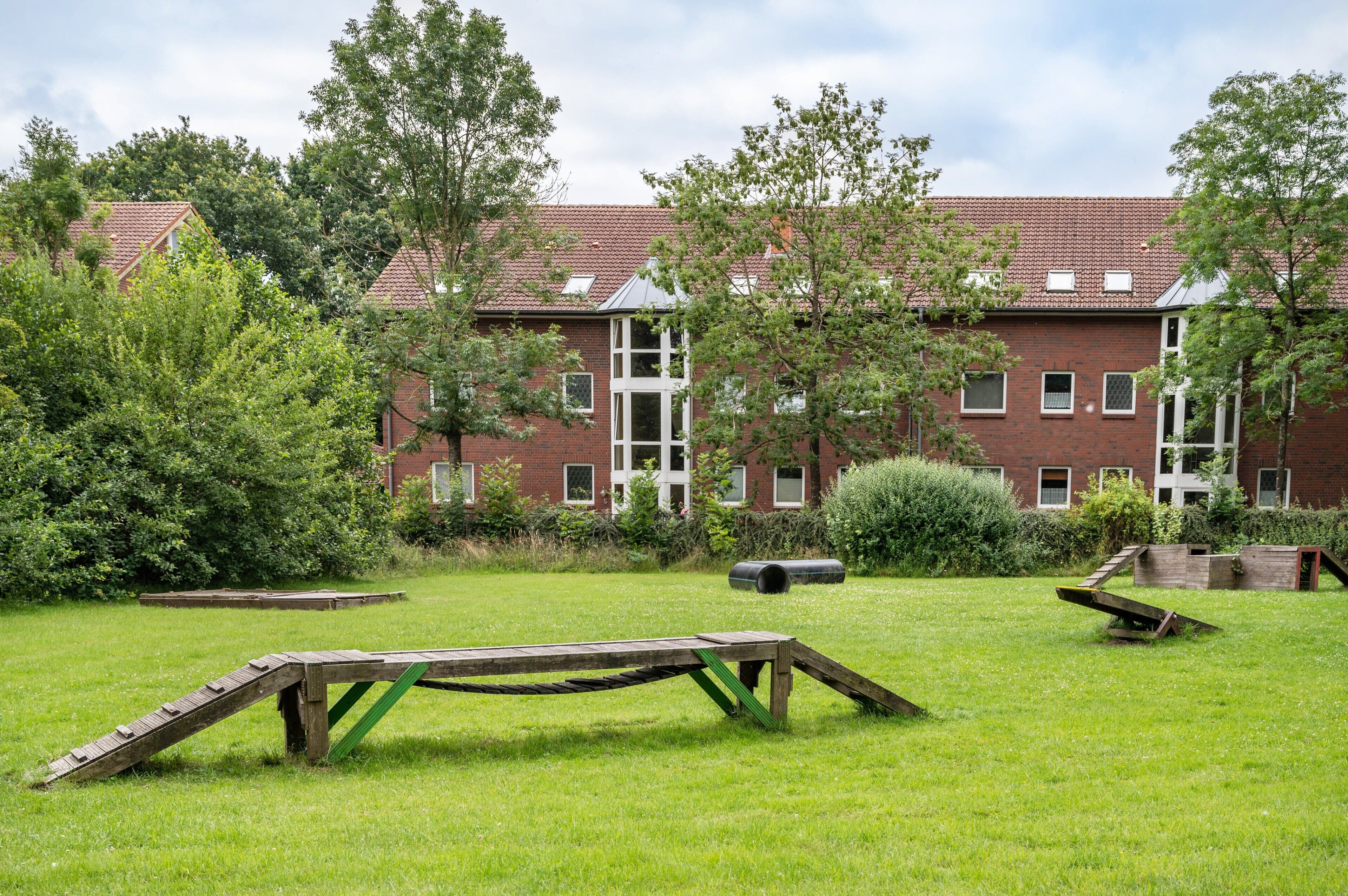 A grassy outdoor area with a wooden structure, trees, and a brick building with windows.