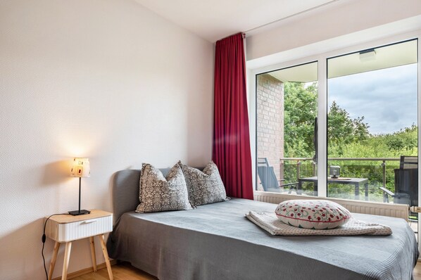 A bedroom with a bed, a bedside table with a lamp, a window with a view of greenery, and a balcony.