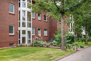 A red brick building with multiple windows, a door, and a small garden with pink flowers.