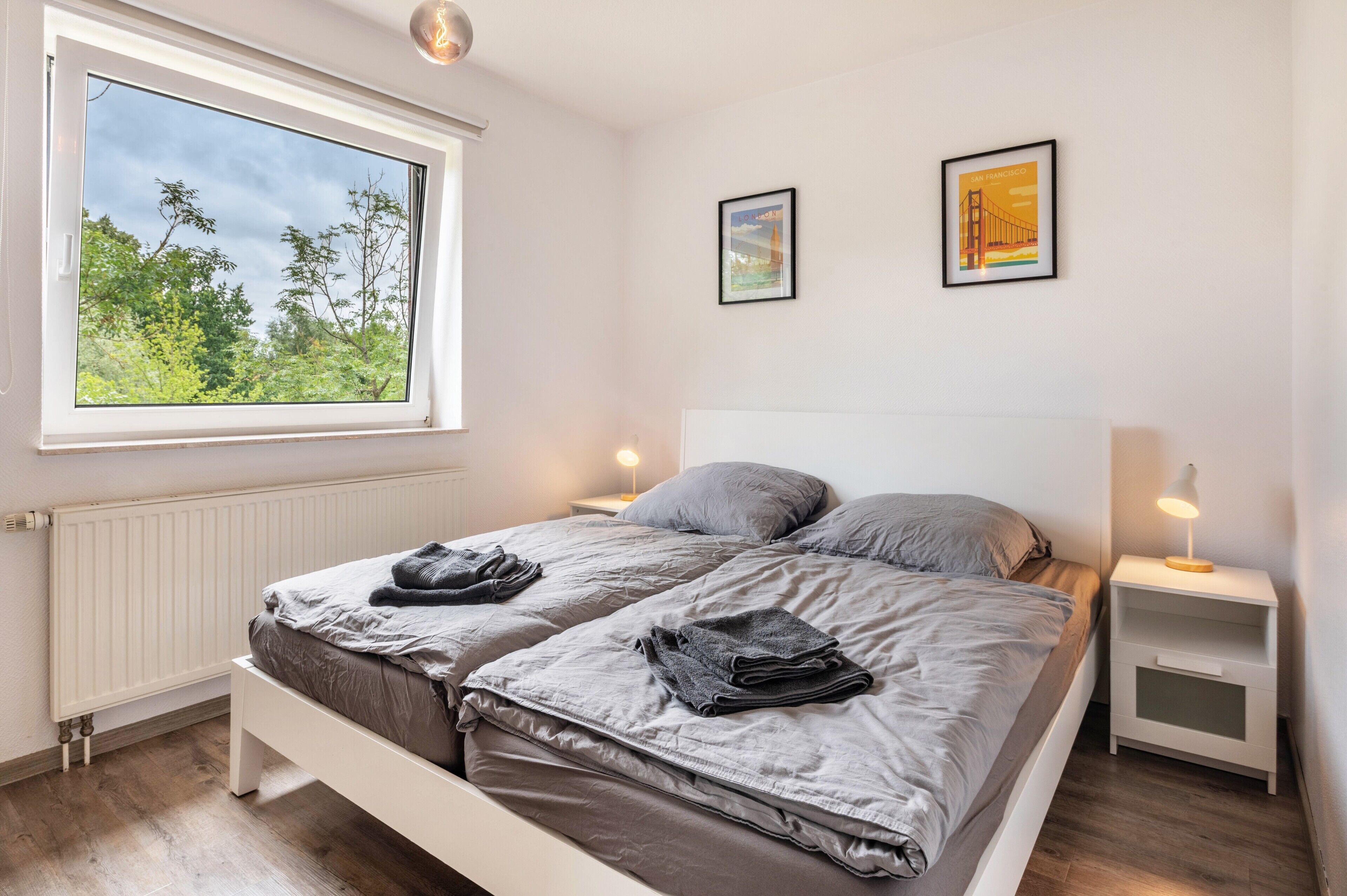 Ein Doppelbett mit grauer Bettwäsche, zwei Nachttische mit Lampen, ein Fenster mit Blick auf Bäume und zwei gerahmte Bilder an der Wand.