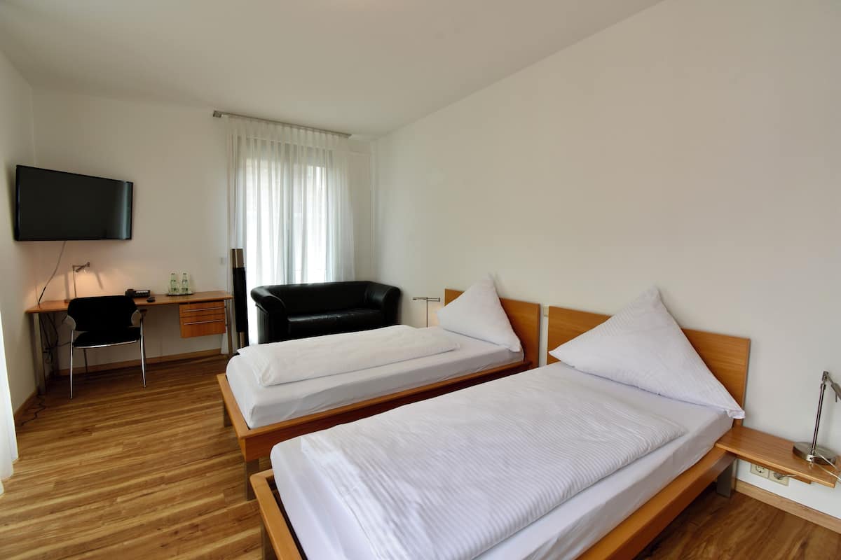 Habitación de hotel con dos camas individuales, un escritorio con silla, televisor montado en la pared y una ventana con cortinas.