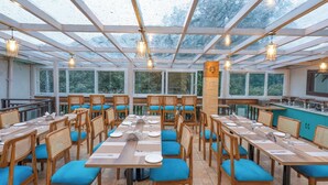 A restaurant with long wooden tables and blue cushioned chairs, large windows, and a glass ceiling. - Excel Hotels & Resorts Mall Road Nainital (Nainital)