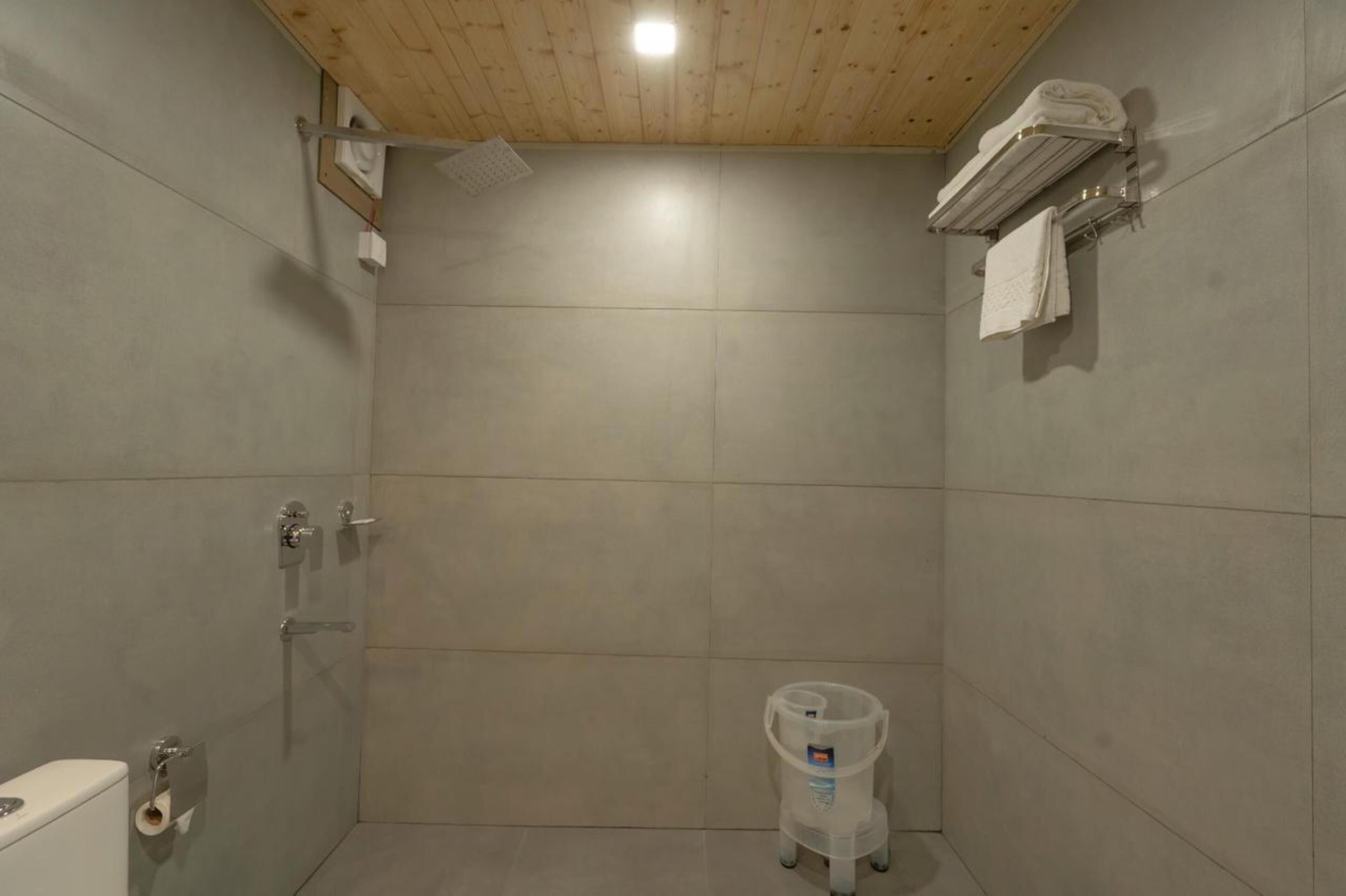 Un pequeño baño revestido con azulejos, con techo de madera, inodoro y un recipiente de plástico para agua.
