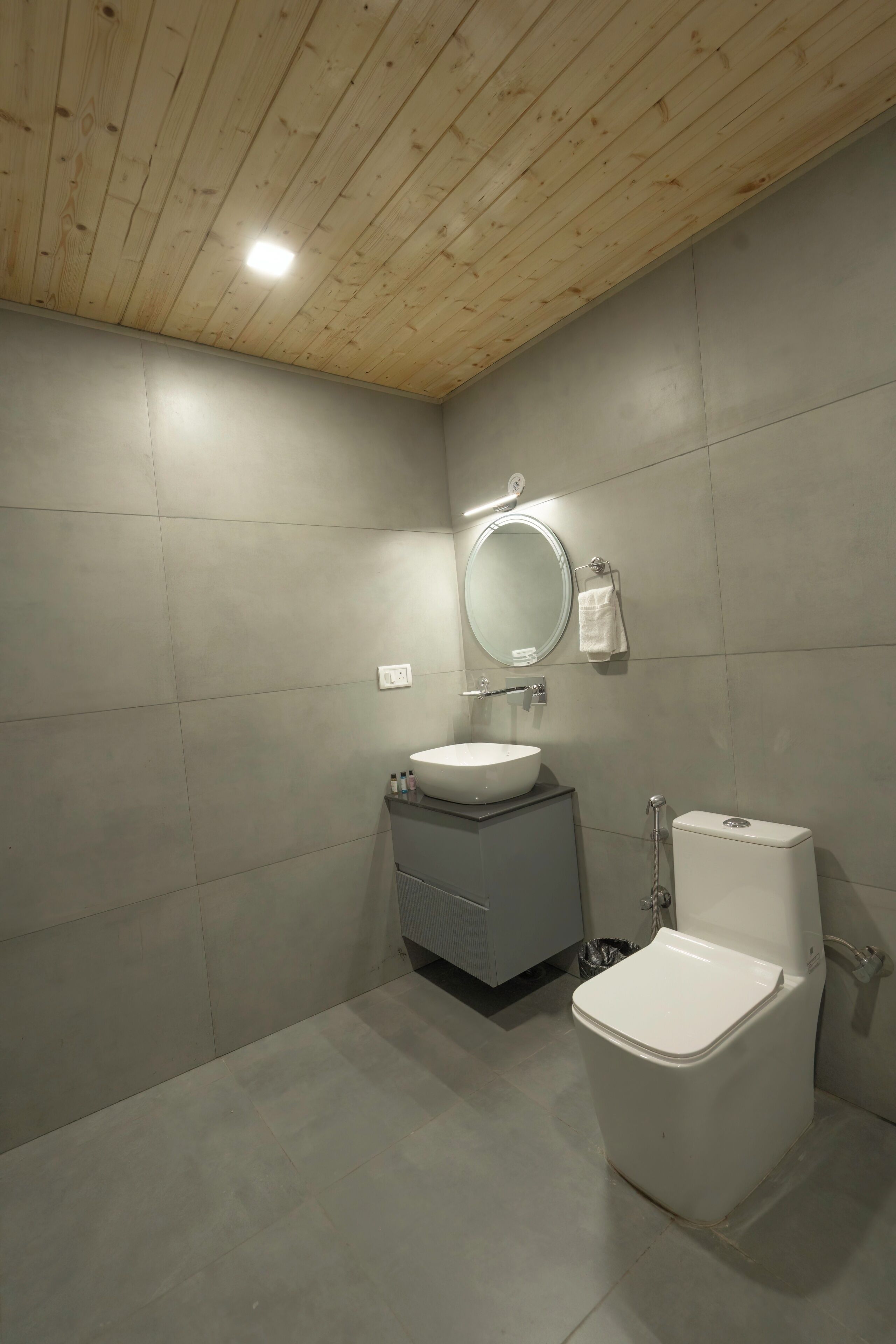 Un baño con techo de madera, un espejo redondo, un lavamanos, un inodoro y un cubo de basura.