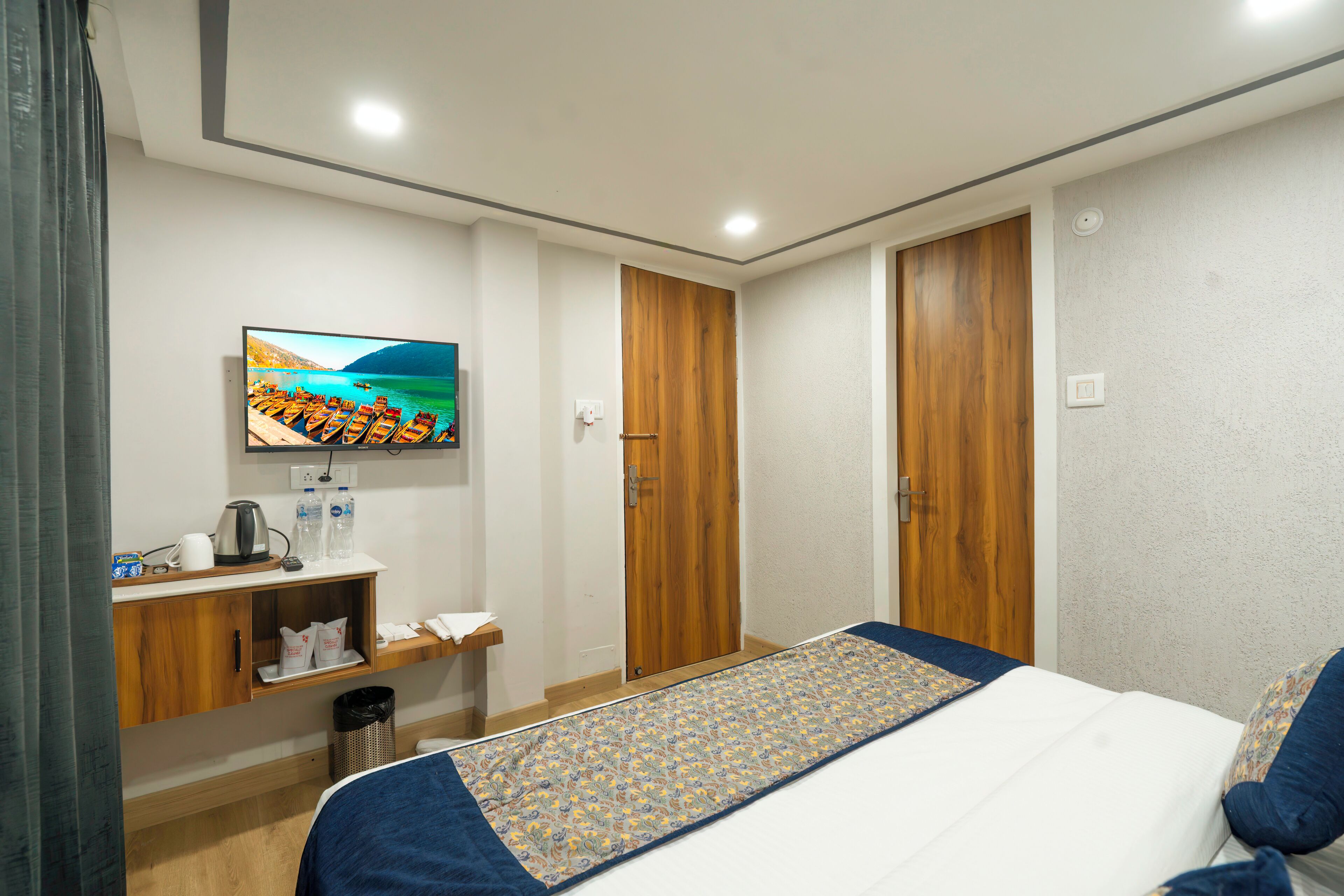 A hotel room with a bed, a TV mounted on the wall, a wooden door, and a small shelf with items.