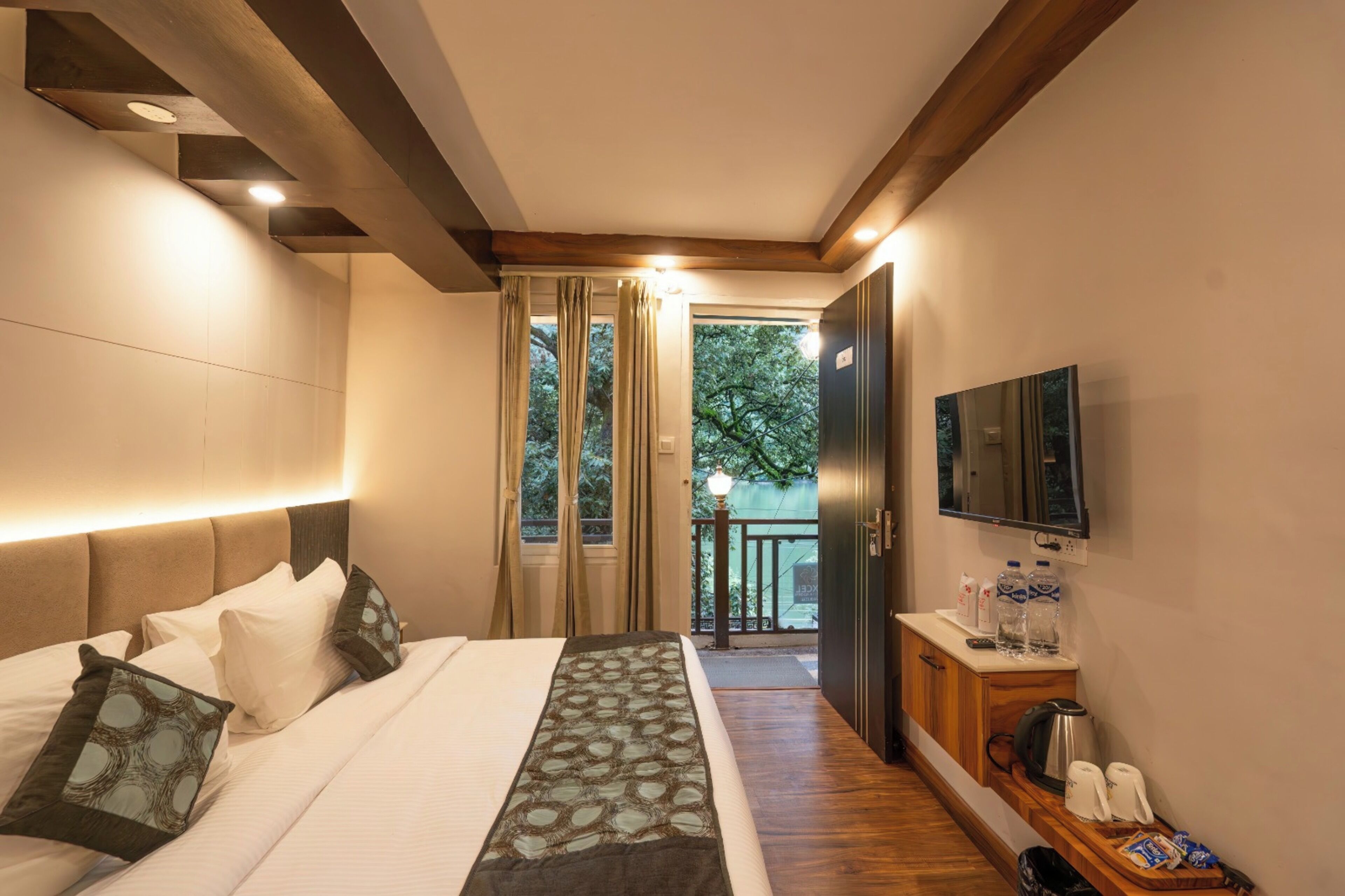 A modern hotel room with a large bed, a TV mounted on the wall, and a balcony with a view of trees.