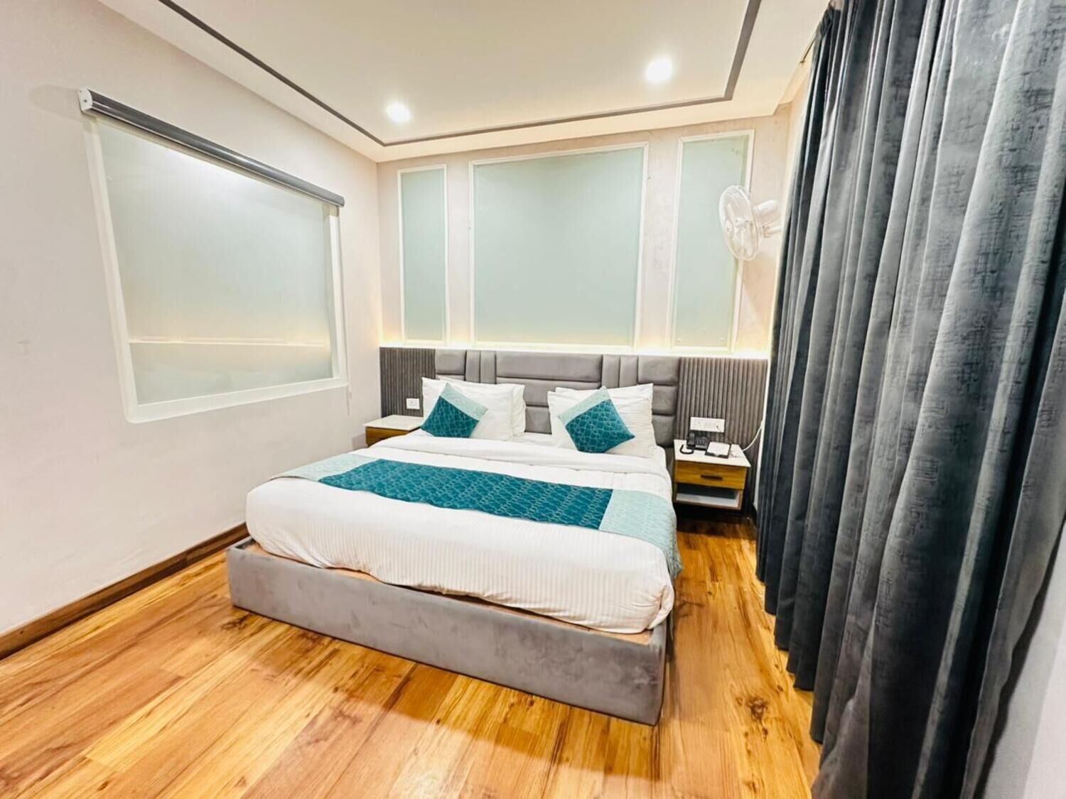 A modern hotel room with a large bed, a bedside table, a nightstand, a window with blinds, and a curtain.