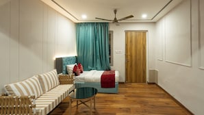 A modern room with a sofa, a small table, a bed with teal curtains, a ceiling fan, and a wooden door. - Excel Hotels & Resorts Mall Road Nainital (Nainital)