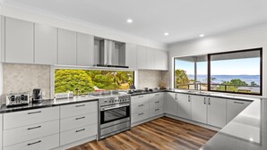A modern kitchen with white cabinets, a stainless steel stove, and a large window offering a view of greenery and a body of water. - Beach Road Bliss in Rhyll (Rhyll)