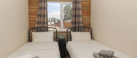 A bedroom with two beds, wooden panel walls, a window with curtains, and a view of an outdoor area.