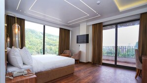 A hotel room with a large bed, a flat-screen TV, a wooden desk with a telephone, and a balcony with a view of greenery. - Hotel Malsi Mist (Dehradun)
