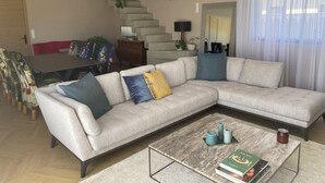 A living room with a sectional sofa, a coffee table, a dining table with chairs, a staircase, and a large mirror.