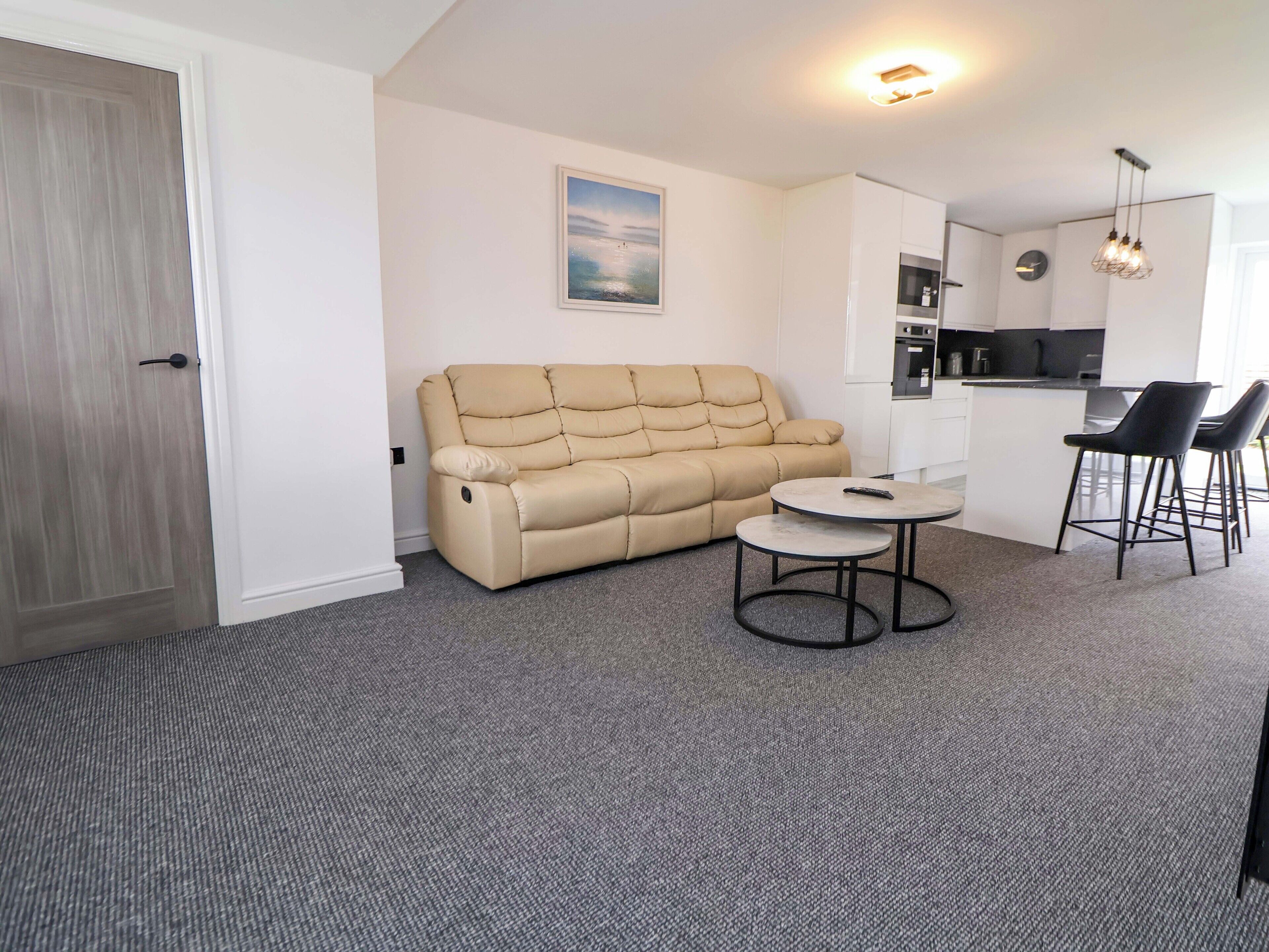 Een moderne woonkamer met een beige bank, een klein rond tafeltje en een keukenhoek met barkrukken.