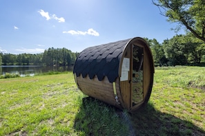 A wooden barrel-shaped structure with a curved roof, situated on grass near a body of water. - Tiny Home 'Mit Blick Auf Den See' with Private Garden, Wi-Fi and Air Conditioning (Fitzen)